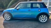 A blue Mini Cooper S with a white roof and side mirrors is parked in a driveway in front of a house. The car's windows are slightly reflective, with a rainbow lens flare effect partially visible on its side. The background includes part of a garage, bushes, and neighboring houses.