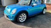 A shiny blue 2004 Mini Cooper S with a black roof and white side mirrors is parked in a driveway. The R53 model boasts a sporty design with silver alloy wheels and the classic Mini Cooper emblem on the front grille. Part of a brick building and an open carport are visible in the background.