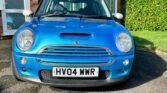 A blue 2004 Mini Cooper S with white side mirrors is parked on a driveway in front of a red-brick house with white window frames. The car's license plate reads "HV04 WWR." A well-maintained green hedge surrounds the lawn to the right side of the house. The sky is clear and blue.