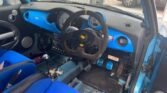 Interior view of a sports car showing a modified dashboard and steering wheel. The dashboard features blue accents, and there are two large gauges behind the suede-wrapped, GTR-branded steering wheel. The MINI-inspired design includes blue racing seats, a fire extinguisher on the floor, and exposed foot pedals.