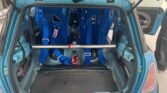 A blue MINI with its rear hatch open reveals a racing setup. The interior includes two blue racing bucket seats with harnesses, a roll bar marked "GTT," and a fire extinguisher mounted on the floor. The rear seats and much of the interior trim have been removed, exposing the metal framework.