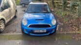 A blue Mini Cooper S with the license plate "RO56 HNH" is parked facing forward at the edge of a road. It is flanked on the left by a silver van. To the right, there is a wooden fence and overgrown greenery. In the background, a dark-colored SUV is parked near a residential area.