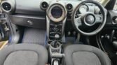 The interior of a MINI car features two front seats, a steering wheel with the iconic Mini logo, and a dashboard with multiple dials and controls. The central console displays a gear stick, cup holders, and buttons. The car has dark fabric seats and floor mats with a blue border.