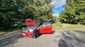 A red MINI with its hood and driver's side door open is parked on a paved area surrounded by tall green trees and foliage. The car is illuminated by bright sunlight, casting distinct shadows on the ground. The background features a combination of greenery and a partly cloudy blue sky.