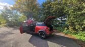 A red MINI compact SUV is parked on a sunlit, leaf-strewn road with its front, rear, and trunk doors open. The scene is framed by green and autumnal trees against a bright blue sky with a few clouds, creating a natural, inviting atmosphere.