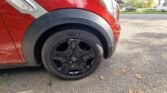 A close-up view of a black wheel with a Michelin tire on a red MINI, parked on a paved surface. The car's front fender and part of the bumper are visible. Fallen leaves are scattered on the ground, and a grassy area with trees is in the background.