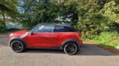 A red two-door compact crossover MINI SUV with black accents and wheels is parked on a paved road. The car features a black roof and side mirrors, situated next to dense green foliage. Dappled sunlight illuminates part of the vehicle and the road, under a clear blue sky visible through the trees.