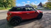 A compact red MINI with a black roof is parked on a paved area in the park. Trees with fall foliage surround the scene. In the background, there's a gazebo with a pointed roof and a picnic table. The sky is blue with scattered clouds, suggesting a crisp, clear autumn day.