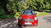 A red MINI Cooper with the registration number ND13 HFN is parked on a tree-lined road. The car is facing the camera, set against a backdrop of lush green trees and a partly cloudy blue sky. Fallen leaves are scattered on the paved surface, indicating an autumn setting.