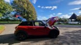 A red MINI hatchback with both its hood and rear hatch open is parked on a paved area near a large grassy field and several tall trees. In the background, there is a small, gazebo-like structure. The sky is blue with scattered clouds, suggesting a sunny day.