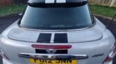 A silver MINI Cooper S is parked in a residential area, flaunting a black racing stripe that runs down the center of the back and roof. The tailgate license plate reads "FG12 JNN." The photo captures the car from behind, with a grassy area and wooden fence visible in the background.