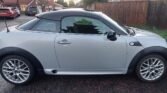 A sleek, silver MINI Coupe with a black roof and shiny alloy wheels is parked on a residential street. Houses and a wooden fence are in the background. The car boasts black trim around the wheel arches and side skirts. The road surface appears slightly wet, possibly after rain.