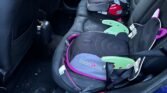 Backseat of a MINI car showing two children's booster seats. One seat with a red base and black cover is positioned on the left, while another seat with a black cover, pink trim, and label "Boost Apak" is on the right. The car interior is black with visible scuff marks and dirt on the floor.