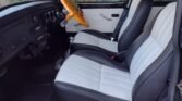 Interior of a classic MINI featuring a wooden steering wheel, black dashboard, and two front seats upholstered in white leather with black trim. The door panels are white with black accents. A 4-point harness is visible on the passenger seat. The background shows a wooden fence and greenery.