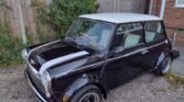 A classic black and white MINI Cooper with a white top is parked on a gravel driveway. The car features front rally lights, distinctive wide wheel arches, and black alloy wheels. A tall wooden fence and some green bushes are in the background alongside a brick wall.