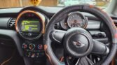 A view from inside a Turbo 2014 MINI Cooper car reveals the steering wheel with the MINI logo, an orange and black cover, and dashboard. The speedometer, fuel gauge, and central screen with car settings are displayed. Yellow fuzzy dice hang from the rearview mirror. The street outside shows parked cars and red brick houses in this EURO6 Mini One D.