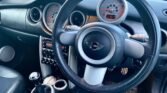 The image shows the interior of a MINI Cooper. The steering wheel, proudly featuring the MINI logo, is prominently in the foreground. The dashboard includes round speedometers and air vents. On the center console, there are various controls and a gear stick, accompanied by cup holders.