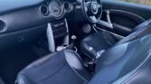 The image shows the interior of a MINI car featuring a black leather steering wheel, a gray dashboard with a large circular speedometer, and black leather seats with gray accents. The center console includes a manual gear shift, an entertainment unit, and various control buttons. A glimpse of a wooden fence is visible through the windows.