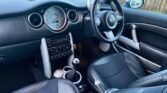 The image shows the interior of a MINI Cooper, featuring a black leather steering wheel with the MINI logo, a large central speedometer, additional round dials, black leather seats, and a manual gear stick with a shiny knob. The dashboard and panels are designed with metallic and black accents.