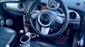         Interior of a car featuring a black steering wheel with a MINI logo, a speedometer and tachometer, controls for audio and climate, and a gear stick. The dashboard and seats are primarily black with silver accents, while the floor sports striped metal pedals and a black mat.