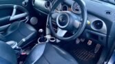 Interior of a car featuring a black leather driver's seat with silver and black accents on the door and dashboard. The steering wheel prominently displays the MINI logo. The dashboard includes a large speedometer and other gauges. There are three pedals, a manual gear stick, and two cup holders. Exterior is partially visible.