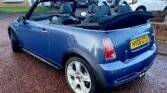 A blue MINI convertible with its soft top down is parked on a wet, red gravel driveway. The vehicle features white alloy wheels and a UK registration plate reading "HY06 GTU." Several houses and other cars are visible in the background, indicating a residential area.