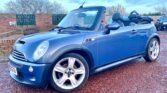 A blue MINI Cooper convertible is parked outdoors on a red gravel surface. The car has its top down, revealing a view of the black interior. The vehicle features silver alloy wheels, a black hood scoop, and a UK license plate. A brick wall and trees are visible in the background.