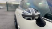 Close-up of a side mirror on a white MINI, displaying a black-and-white Union Jack design. The background is slightly blurred, with hints of a fenced area and other parked cars under a cloudy sky, emphasizing the mirror's distinctive design. The focus is on the side mirror.