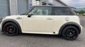 A white MINI Cooper with a black roof and black alloy wheels is parked in front of an industrial building with a grey exterior and multiple garage doors. The car features red brake calipers and a chrome fuel cap on its rear side, set against a paved surface and slightly cloudy sky.