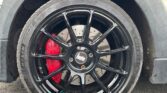 Close-up of a car's front wheel with a black multi-spoke rim, branded "Borbet," and a visible red brake caliper. The cream-colored MINI's body above the wheel features a sleek black fender. The pavement and part of a building in the background are slightly blurred.