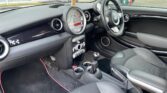The interior of a modern MINI car, featuring a black and silver color scheme with leather seats. Visible are the steering wheel, dashboard with a large central speedometer, gear shift, and various controls. The overall design is sleek and compact, with red accents on the floor mats.