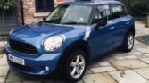 A blue MINI Countryman is parked on a stone-paved driveway in front of a brick house with gray window shutters. The house has large windows with black frames. Trees and shrubs are visible in the background. The vehicle's license plate reads "WM63 OZD".