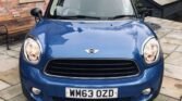 A blue MINI Cooper, with the license plate "WM63 OZD," is parked on a tiled driveway next to a brick house. The background includes a wooden shed and leafless trees. The car's front view prominently displays its headlights, grille, and iconic MINI emblem.