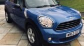 A blue MINI Cooper sits parked on a beige stone driveway, surrounded by a grassy yard and trees. The car's front is angled towards the camera, showcasing its headlights, grille, and silver alloy wheels. Its UK license plate reads "WM63 OZD." A small shed is visible in the background.