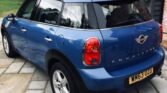 A blue MINI Countryman is parked on a stone-paved driveway outside a house with brick and wooden exterior walls. The car features distinctive red taillights, white trim around the wheel arches, and a black roof. Trees and greenery are visible in the background. The license plate reads "WM63 OZD.