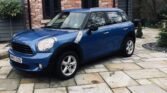 A blue MINI Cooper SUV is parked on a stone-paved driveway in front of a brick and wood-paneled house. The surrounding area is landscaped with shrubbery and trees. The vehicle, which has a sunroof, is positioned at an angle, with its front facing left. The license plate reads "M663 OZO.