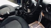 Interior view of a blue MINI featuring black fabric seats, a steering wheel with the iconic MINI logo, a manual gear shift, and cup holders between the seats. The driver’s door is open, revealing a paper floor mat adorned with blue footprint designs on the driver's side.