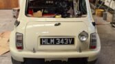 A classic Mini Cooper with license plate HLM 343V is parked in a cluttered garage. The rear window is missing, revealing the car's vintage steering wheel and dashboard. Various tools and car parts are scattered around, including a cardboard box and a container on the floor, evoking pure automotive nostalgia.