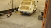 A classic Mini in a white garage is covered in masking tape, prepping for painting. The vehicle is raised on jack stands, with plastic sheeting on windows and lights. The garage has concrete floors, a broom, a car jack, and various items including a tire and crates scattered around.