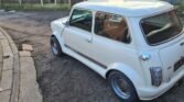 A classic white Mini Cooper, featuring black racing stripes on the side and silver alloy wheels, is parked on a gravel and dirt area beside a curved concrete path. Facing away from a wooden fence and gate, its driver's side visible in the photo, this classic Mini exudes timeless charm.