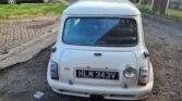 A classic white Mini, likely an older model, is parked on a rough driveway next to a grassy area and wooden fence. The license plate reads "HLM 343V." The car boasts two rear exhaust pipes and a small round taillight on the left. A dark gray hatchback is parked in the background.