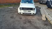 A white classic mini with the registration plate "HLM 343V" is parked on a rough, gravelly surface next to a brick wall. Partially visible to the right are two other vehicles, one silver and one dark-colored, parked on a paved area. The classic car's front grill is mostly black.