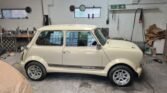 A cream-colored classic MINI 850 car with black stripe decals is parked in a spacious workshop with a concrete floor. The car features rounded headlights, chrome accents, and silver alloy wheels. The workshop includes large windows, various tools, and an emergency exit sign on a white brick wall.