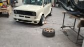 A white vintage classic Mini with a modified front bumper and wide tires is parked in a cluttered garage workshop. The area has various tools, spare parts, a wheel on the floor, and workbenches. A blue cabinet and an open door leading to another part of the workshop are visible in the background.