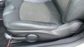 Close-up of a MINI car's driver's seat featuring a black leather and fabric combination. The seat adjustment knob is visible on the side, and the lower part of the center console can be seen in the background. The floor area is carpeted with hints of red wiring partially visible.
