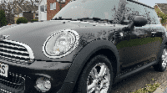 A black MINI Cooper is parked on a street in front of a row of houses. The car has a sleek, modern design with silver alloy wheels and a chrome-trimmed front grille. The overcast sky casts a muted light over the scene, emphasizing the MINI's glossy finish.