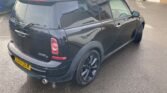 A black MINI Cooper D Clubman with tinted windows is parked on a wet asphalt surface near a building with large windows. The car, perfect for any MINI Clubman enthusiast considering repair or looking for spares, has a visible rear license plate, roof rack, and black alloy wheels. The rear wiper is also visible on the tailgate as sunlight casts gentle reflections on the vehicle.