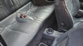 Interior view of a Mini Cooper S, showing the backseat and part of the front passenger seat. The backseat is made of black leather and features two headrests with a central armrest folded back. The carpeted floor complements the sleek design, while two cup holders are visible for added convenience.