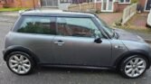 A gray Mini Cooper S R53 is parked on a residential street in front of a row of brick houses with driveways. The car has a black roof and silver alloy wheels. The street and car appear wet, suggesting recent rain. Some small yellow leaves are scattered on the ground.