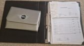 A black binder filled with organized documents and a silver envelope with a MINI logo lies open on a beige carpet. The binder's right side displays multiple invoices and paperwork secured by ring binders, while the left side features the MINI-branded envelope, likely containing additional documents about the 2002 Mini Cooper S.